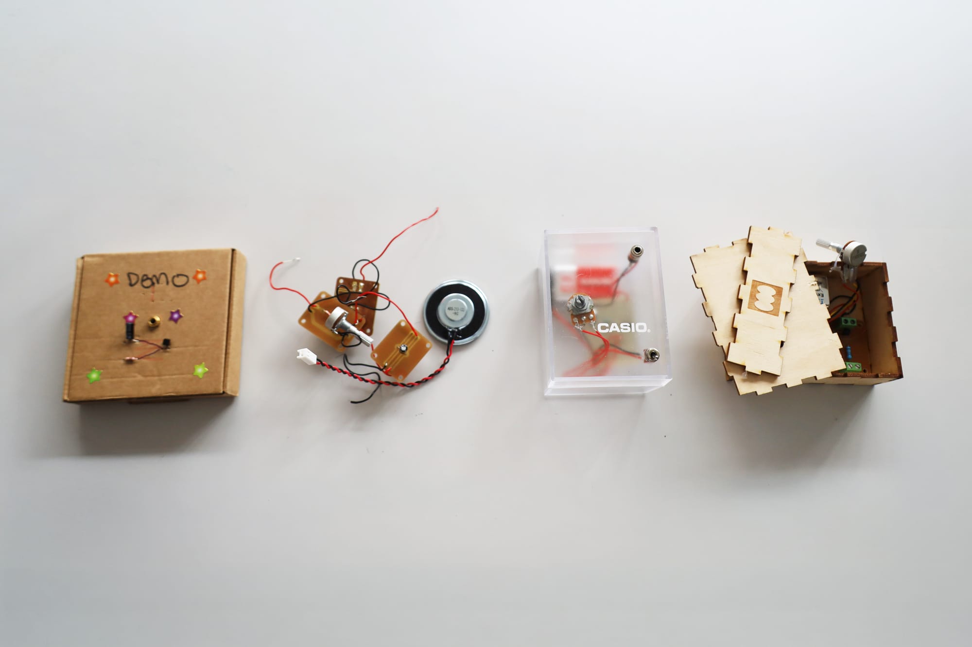 Four analogue synth prototypes sit in consecutive order from earliest (2022) to newest (2026). The first synth is a circuit inside a small cardboard box, the second is a circuit without a board, the third is a small, clear, repurposed plastic Casio box, and finally a custom-build, small, wooden box synth with a laser-cut Synthesize_Her embellishment.
