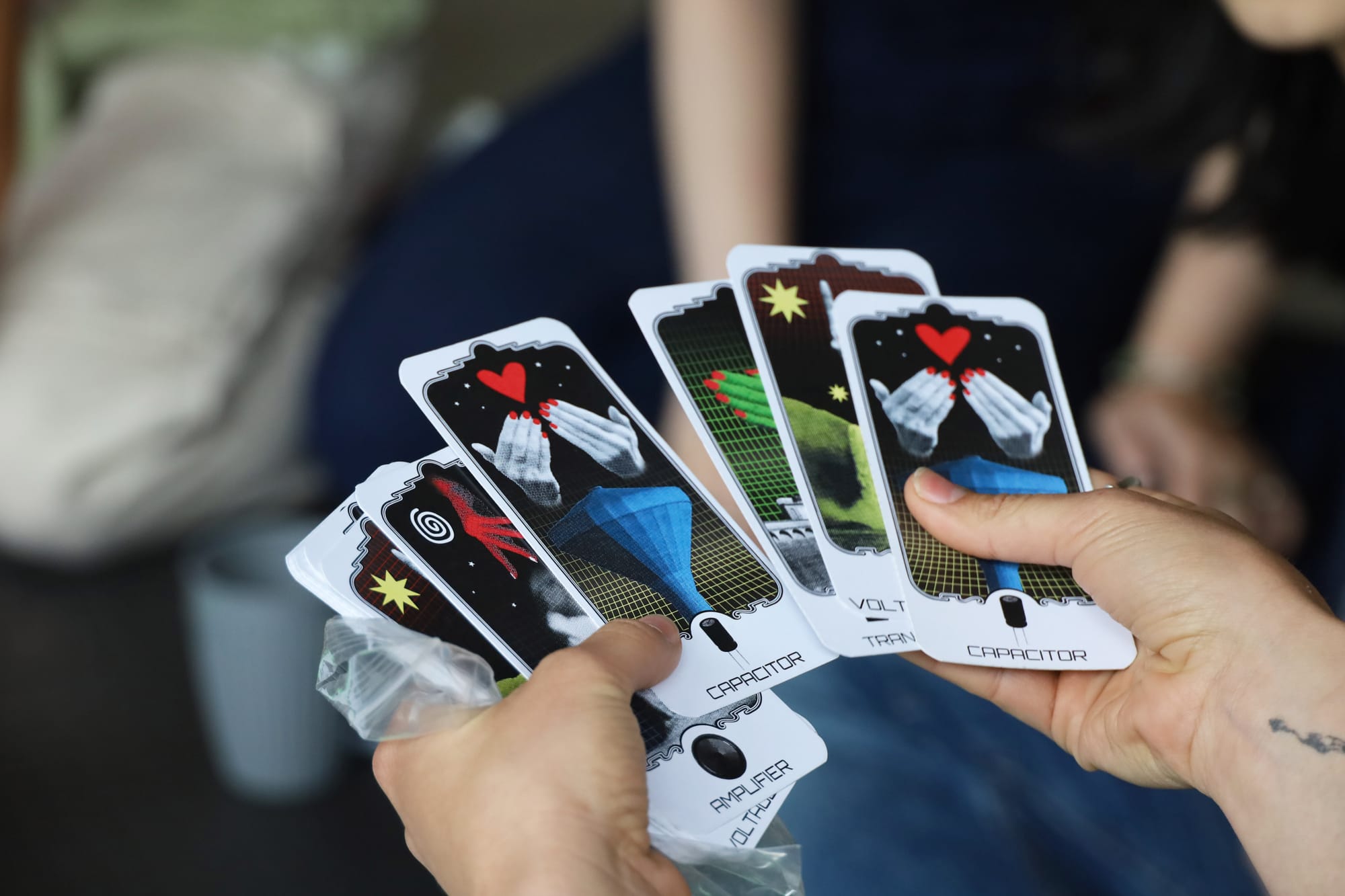 A pair of light-skinned hands hold 5 tarot cards with illustrations of different electrical components - voltage source, amplifier, capacitor, transistor, and resistor.