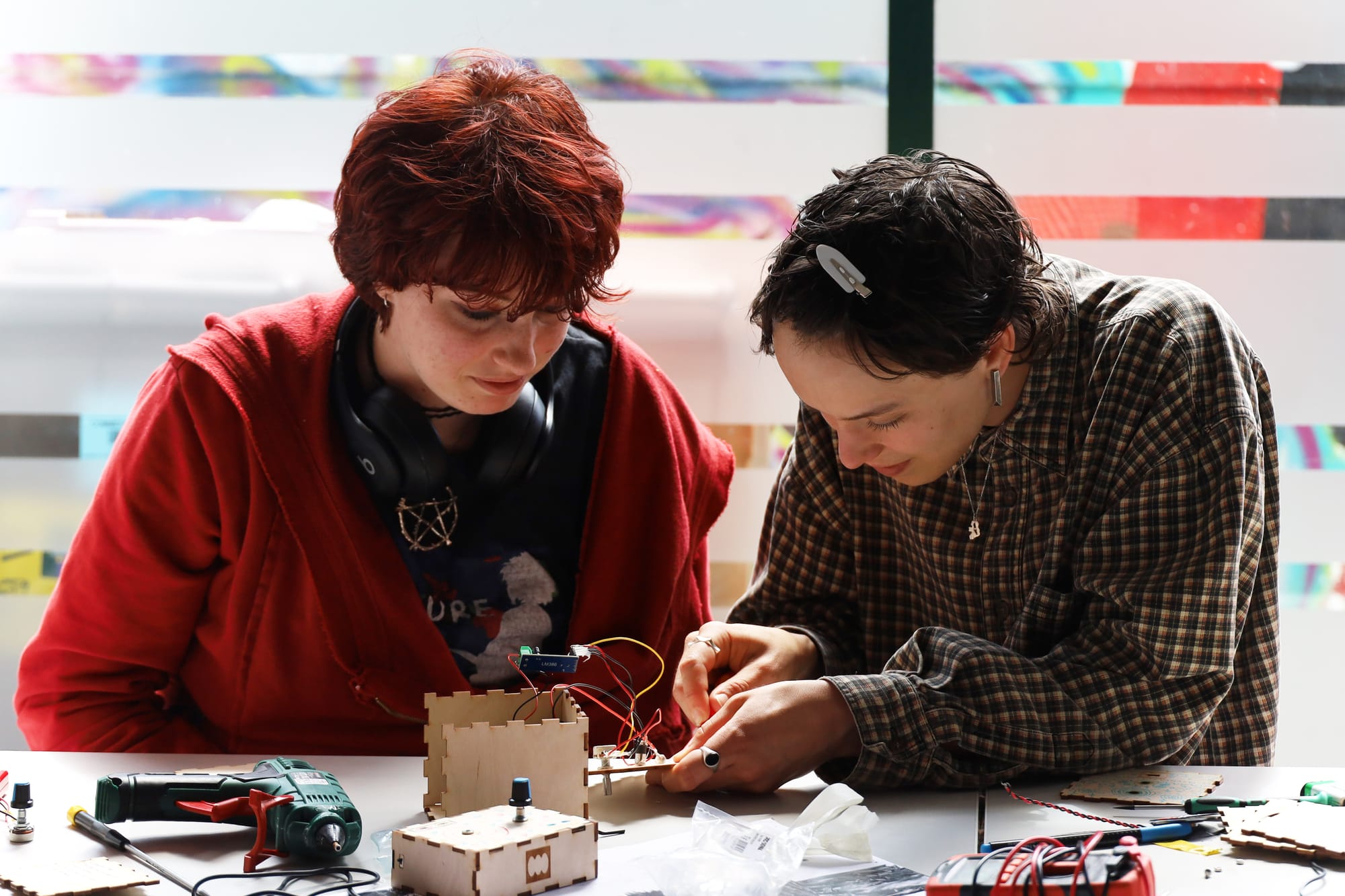 Two alternatively dressed people sit at a long white table with parts of a synth and speaker build in front of them. The person on the left is young with both red hair and a red hoodie. The facilitator, Viva, is on the right wearing a checked shirt with short dark hair. They are checking if the circuit on the partial speaker build is correct.
