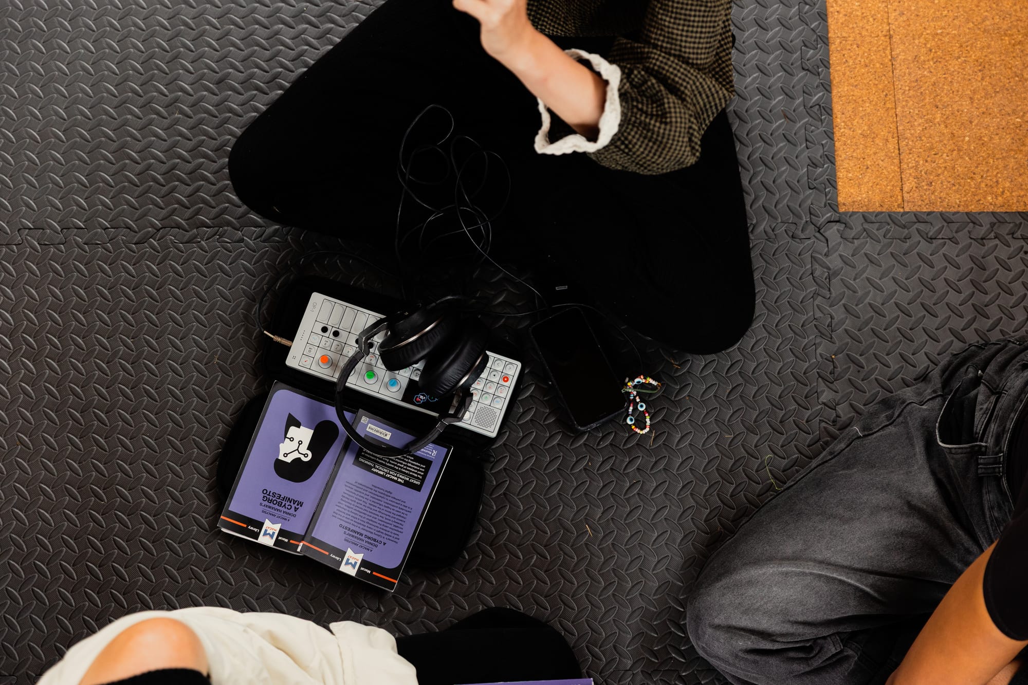 A person in black trousers sits crossed-legged in front of a Teenage Engineering OP-1 synth, and a small text of A Cyborg Manifesto by Donna Haraway.