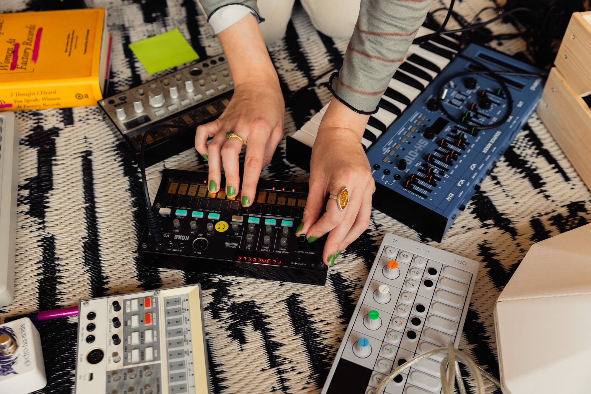 A pair of hands with fair skin, wearing a few rings, plays with a KORG Volca Beats. There are other synths scattered around it on the floor.